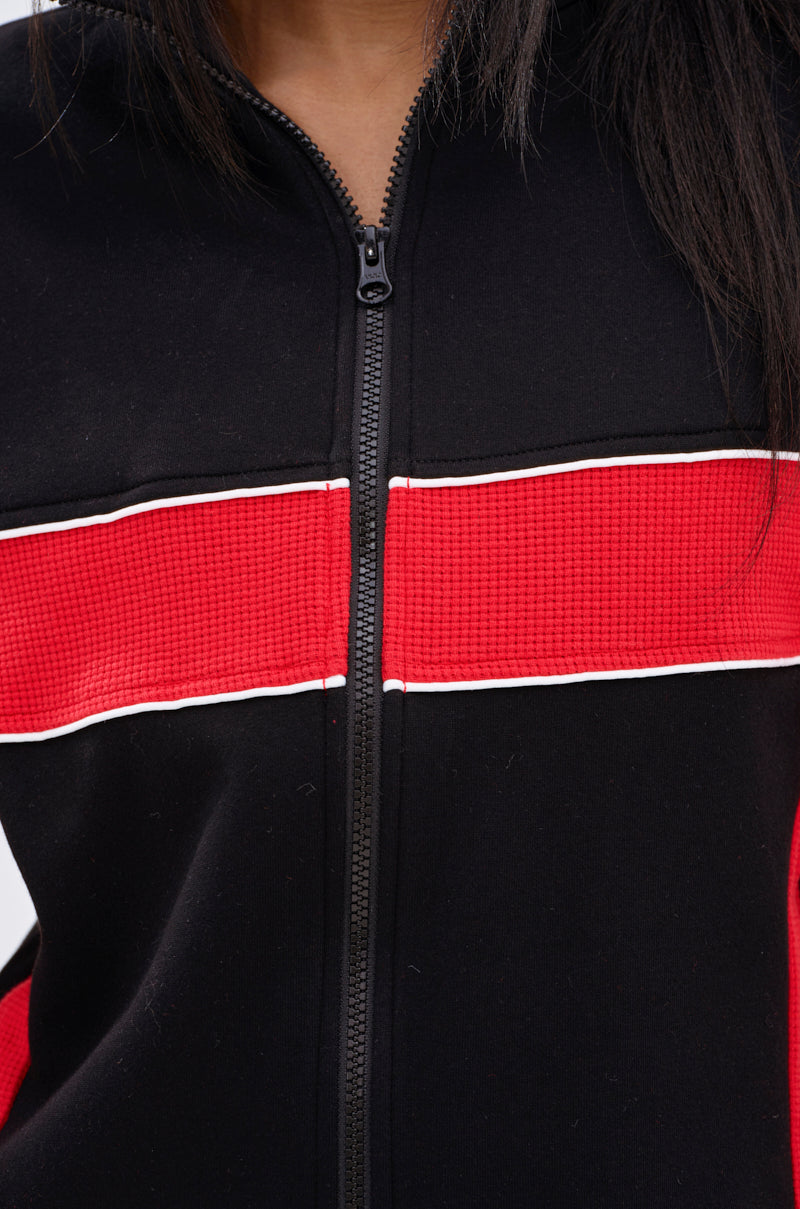 A person wears the LET IT HAPPEN ZIP UP SWEATSHIRT, featuring bold colorblocking with a horizontal red stripe across the chest, outlined by thin white piping. The black sweatshirt has a centered zipper and cropped fit for a modern look.