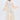 A woman with long gray hair stands confidently against a white background wearing the TAY TAY TROUSER, a cream blazer with sheer, ruffled sleeves, a flowing pleated ivory skirt, gold necklace, and heels.