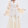 A woman with long gray hair stands confidently against a white background wearing the TAY TAY TROUSER, a cream blazer with sheer, ruffled sleeves, a flowing pleated ivory skirt, gold necklace, and heels.