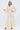 A stylish older woman with long gray hair confidently models the TAY TAY TROUSER—a flowy, cream wide-leg pant—paired with a cream vest featuring ruffled sheer sleeves, gold shoes, and a statement necklace against a white background.