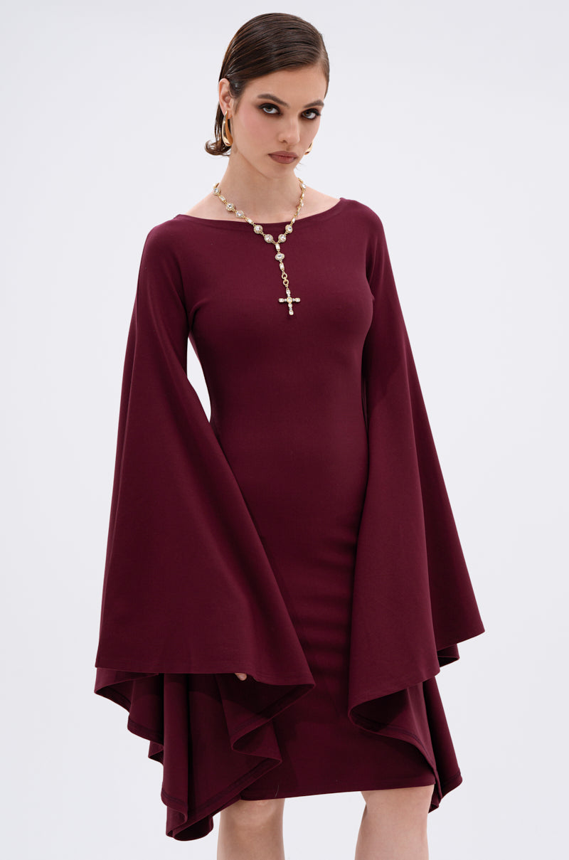 She wears the MEET ME IN THE BALLROOM MIDI DRESS IN BURGUNDY with dramatic bell sleeves and a gold cross necklace, her hair slicked back as she poses against a plain background—an elegant choice for any formal event.