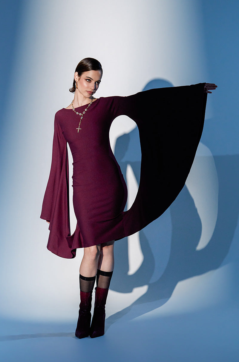 Wearing the MEET ME IN THE BALLROOM MIDI DRESS IN BURGUNDY, a woman poses confidently against a blue-lit backdrop. The fitted midi dress with dramatic asymmetrical long sleeves is styled with a gold cross necklace and matching burgundy boots—formal and striking.