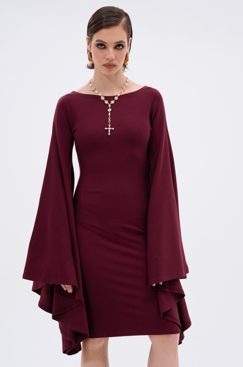 A woman in the MEET ME IN THE BALLROOM MIDI DRESS IN BURGUNDY, featuring dramatic bell sleeves, stands against a white background. Her short, slicked-back hair and pearl necklace with a cross pendant complete her formal look.