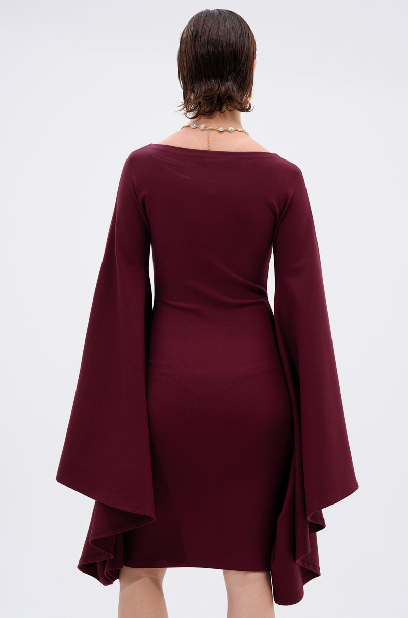 A person with short, dark hair stands against a light gray background wearing the MEET ME IN THE BALLROOM MIDI DRESS IN BURGUNDY. This fitted, knee-length dress features long draped bell sleeves and is styled with a beaded necklace.