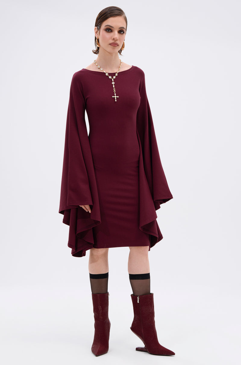 A woman faces forward in the MEET ME IN THE BALLROOM MIDI DRESS IN BURGUNDY with flared long sleeves, perfect for formal events. She styles it with a cross pendant beaded necklace, burgundy boots, and black sheer socks.