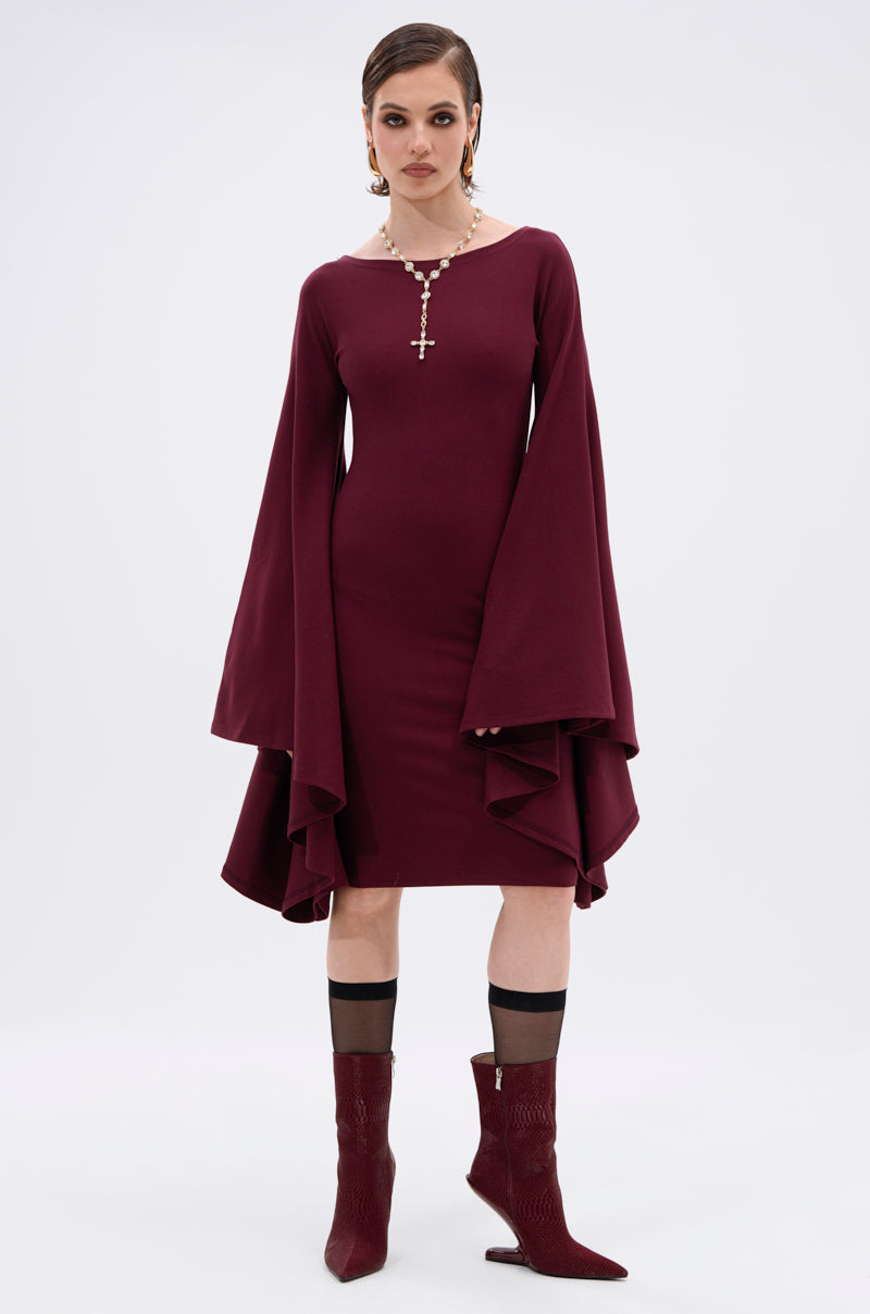 A woman poses against a plain background in the MEET ME IN THE BALLROOM MIDI DRESS IN BURGUNDY, featuring flared long sleeves, maroon boots, a beaded cross necklace, and earrings—ideal for formal occasions.
