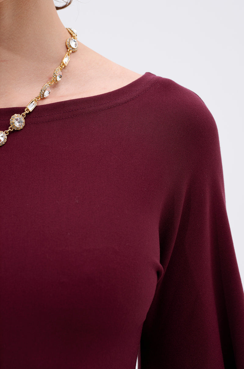 Close-up of MEET ME IN THE BALLROOM MIDI DRESS IN BURGUNDY, featuring a boat neckline—ideal for formal events. A gold necklace with round jeweled accents drapes elegantly around the neck against a plain light background.
