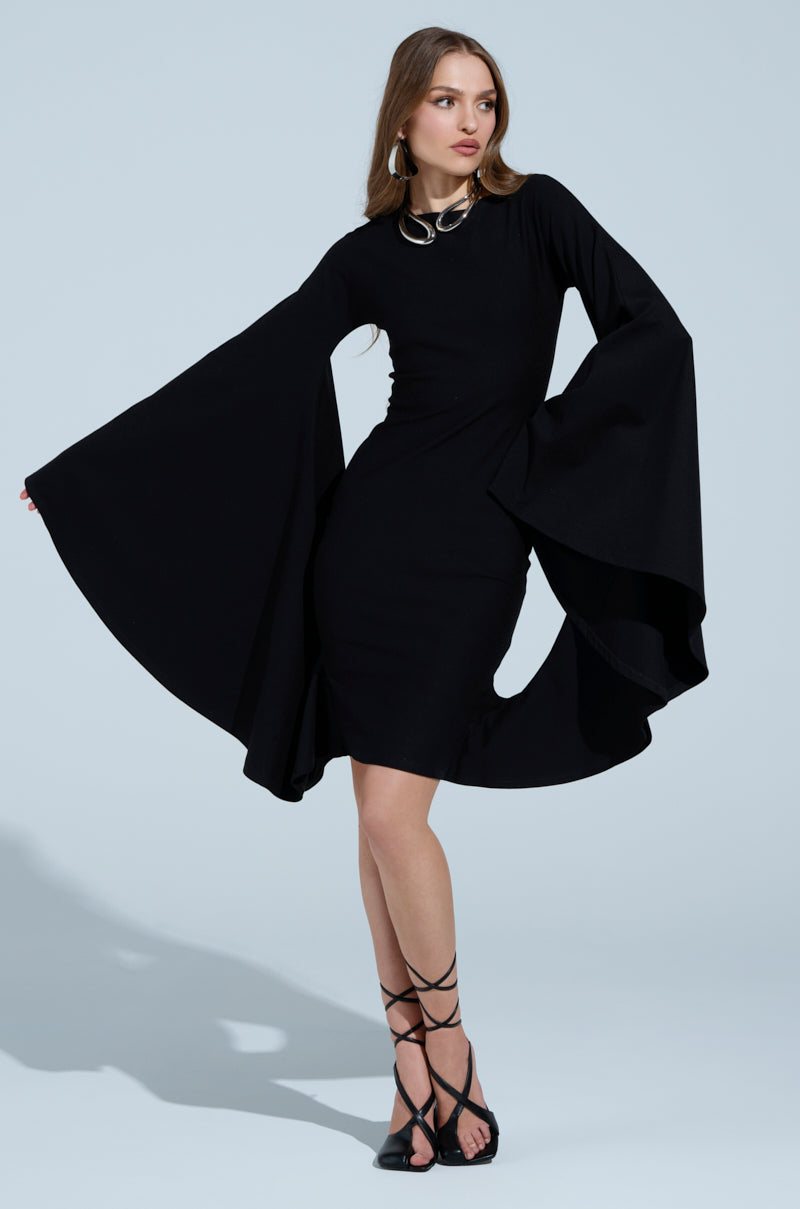 Wearing the MEET ME IN THE BALLROOM MIDI DRESS, a person stands against a light blue background. The black midi dress boasts a boat neckline and dramatic flared sleeves, paired perfectly with strappy black sandals. Long hair cascades down to complete the form-fitting look.