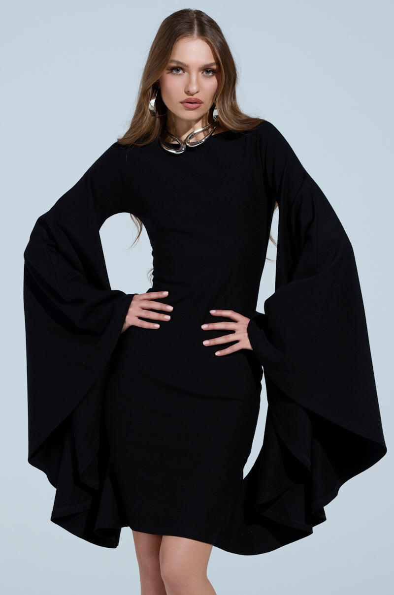 A woman poses confidently in the "MEET ME IN THE BALLROOM MIDI DRESS," featuring dramatic bell sleeves and a boat neckline. She accessorizes with a statement metallic necklace, and her long brown hair contrasts against the solid light blue background.