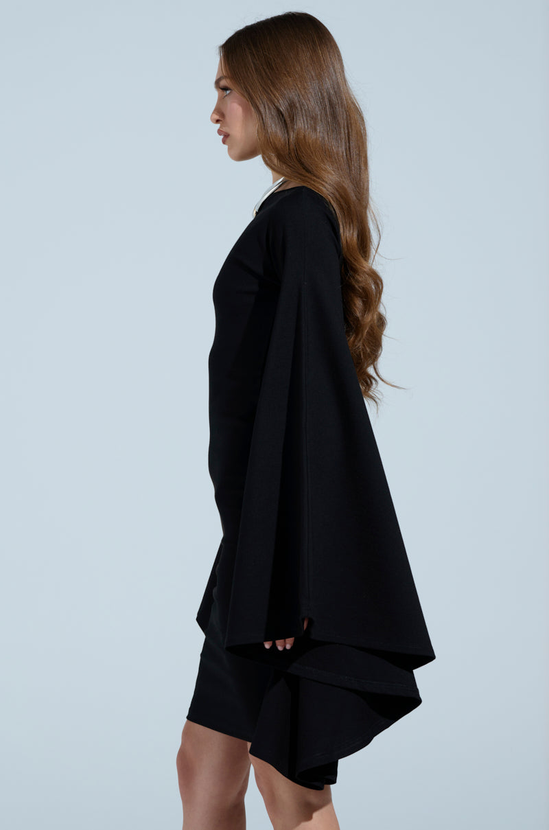 A person with long brown hair is wearing the MEET ME IN THE BALLROOM MIDI DRESS, featuring dramatic bell sleeves and a flowing train. The elegant boat neckline complements their silhouette as they stand in profile against a plain light blue background.