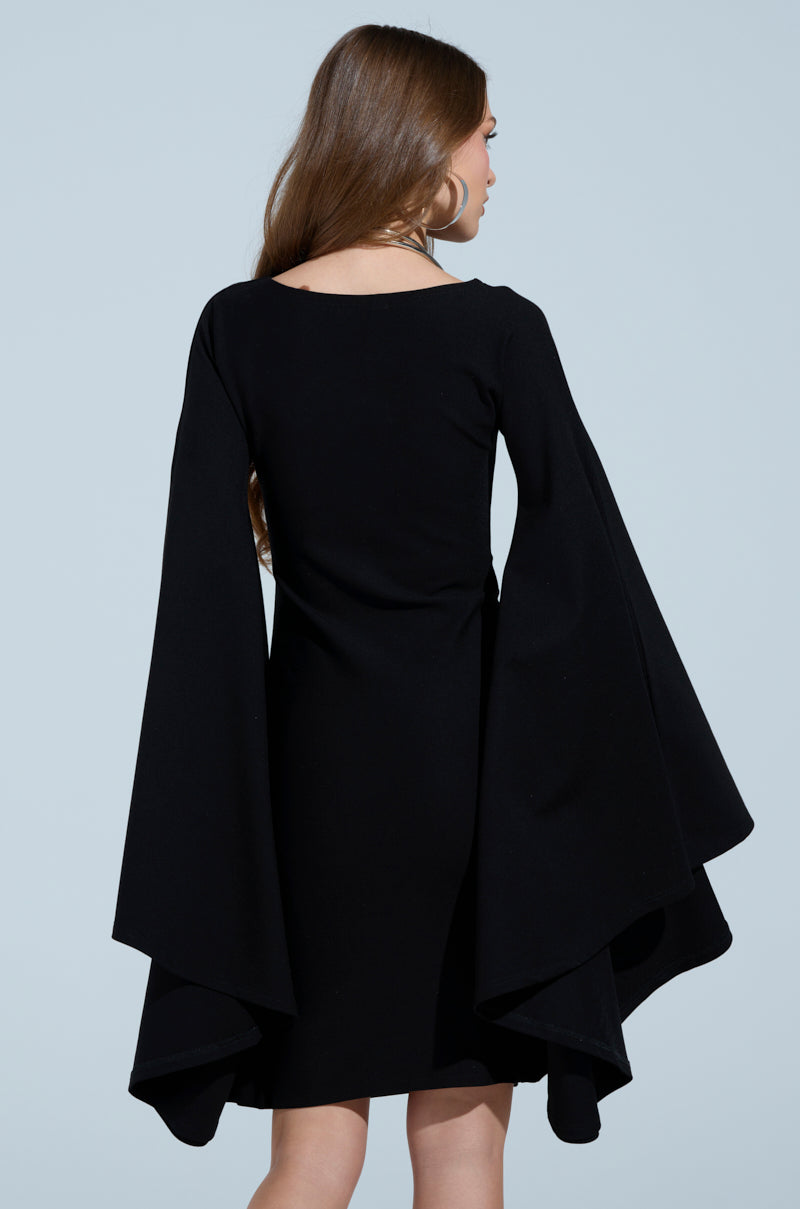 Against a light blue backdrop, a woman with long brown hair is seen wearing the MEET ME IN THE BALLROOM MIDI DRESS, which features striking, flowing bell sleeves. Her back is turned to the camera.
