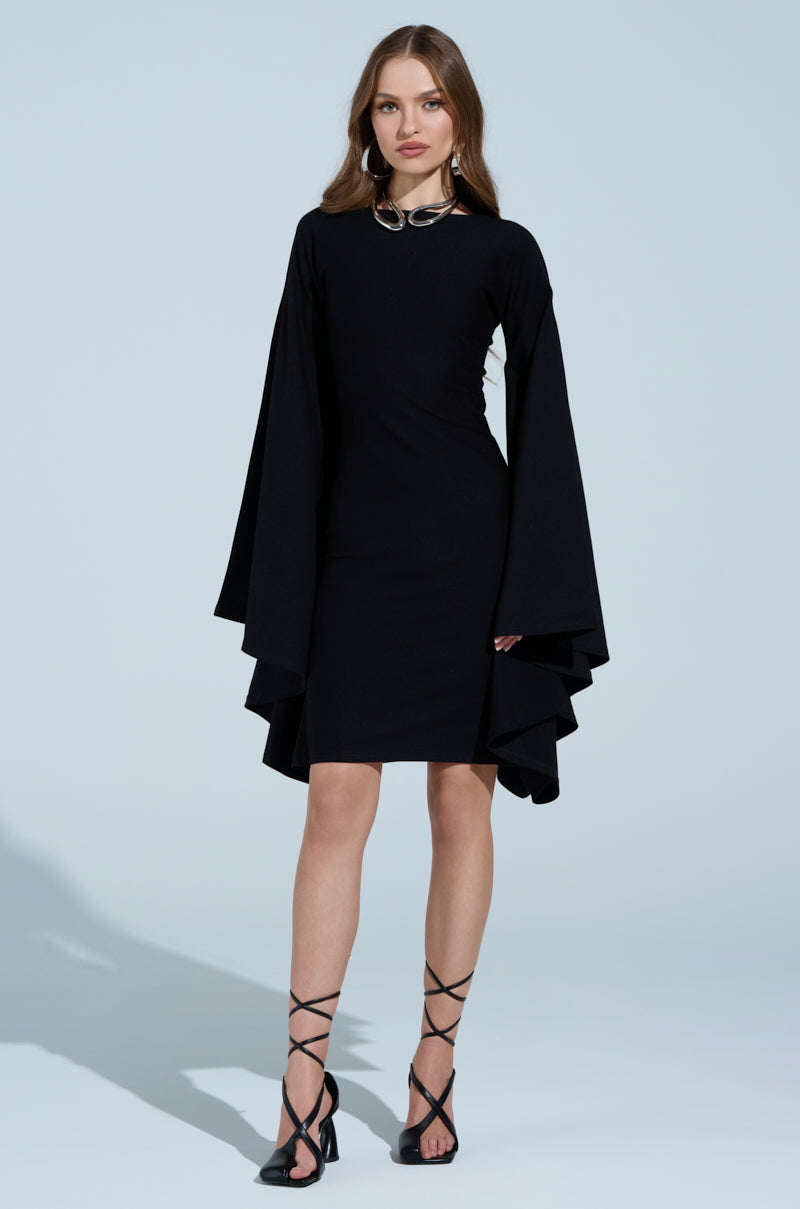 A woman poses confidently against a light blue background wearing the MEET ME IN THE BALLROOM MIDI DRESS featuring dramatic cape sleeves, paired with black strappy high heels. Her long hair and minimal makeup complete the look.
