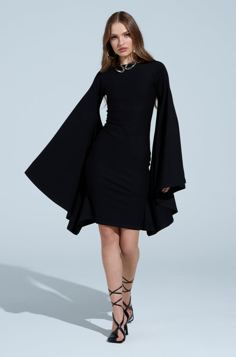 A woman wearing the MEET ME IN THE BALLROOM MIDI DRESS, showcasing its wide boat neckline and flowing bell sleeves, stands facing forward. She pairs it with strappy black heels and her long, straight hair complements the outfit against a light blue background.