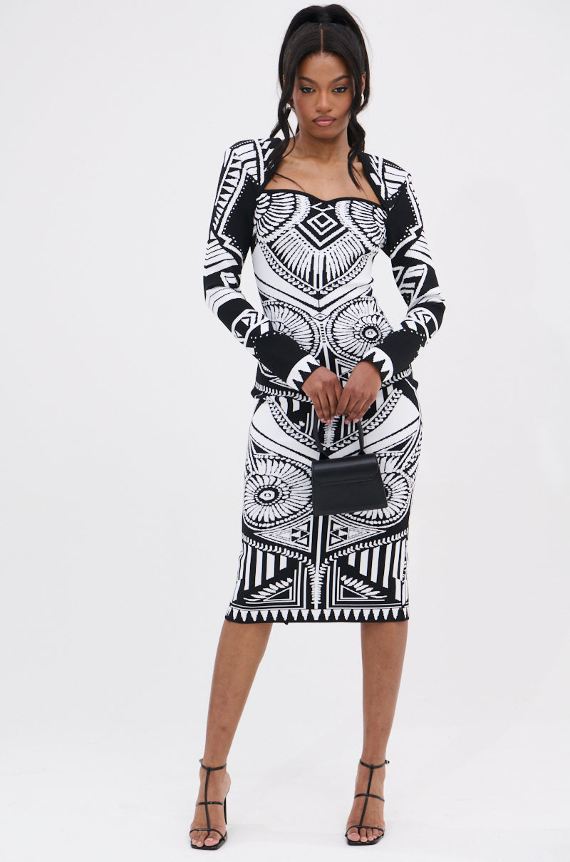 A woman stands against a white background wearing the NOT YOUR AVERAGE WOMAN PRINTED MIDI DRESS with long sleeves, paired with a small black handbag and black strappy heels.