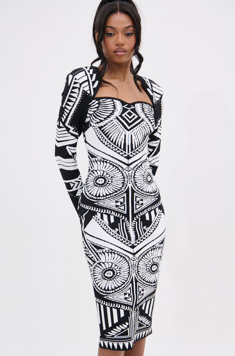 A woman with long dark hair and a neutral expression stands against a white background wearing the NOT YOUR AVERAGE WOMAN PRINTED MIDI DRESS, which features bold black and white geometric and circular patterns.