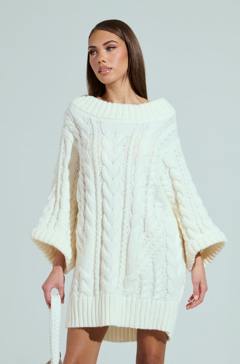 Wearing the LALA LOVE YOU SWEATER DRESS in cream, a person with long, straight brown hair poses against a light gray background. The off-the-shoulder cable-knit dress features wide sleeves and a thick ribbed hem as they hold a small bag in one hand.