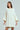 Wearing the LALA LOVE YOU SWEATER DRESS in cream, a person with long, straight brown hair poses against a light gray background. The off-the-shoulder cable-knit dress features wide sleeves and a thick ribbed hem as they hold a small bag in one hand.