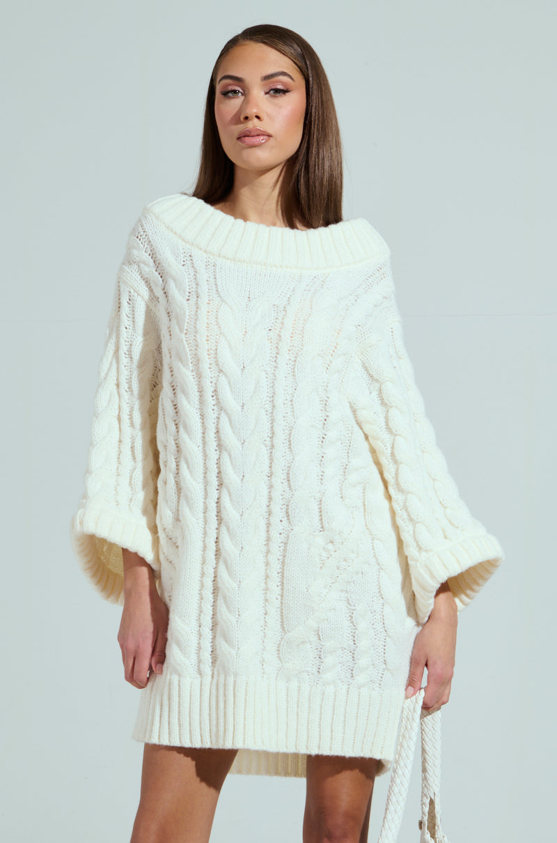 Wearing the LALA LOVE YOU SWEATER DRESS, a person with long brown hair poses against a light gray backdrop. The off-the-shoulder, oversized off-white cable-knit dress showcases wide sleeves and a textured pattern, exuding effortless style with their neutral expression.