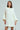 Wearing the LALA LOVE YOU SWEATER DRESS, a person with long brown hair poses against a light gray backdrop. The off-the-shoulder, oversized off-white cable-knit dress showcases wide sleeves and a textured pattern, exuding effortless style with their neutral expression.