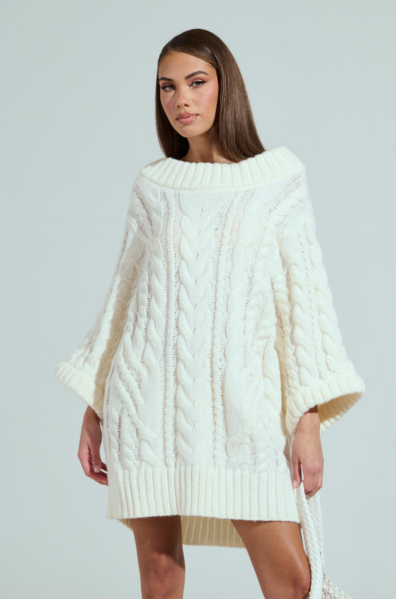 A person wears the LALA LOVE YOU SWEATER DRESS, a chunky, oversized white cable knit outfit with wide three-quarter sleeves and an above-the-knee hem. This stylish piece features an off-the-shoulder design on a plain light gray background.