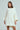 A person wears the LALA LOVE YOU SWEATER DRESS, a chunky, oversized white cable knit outfit with wide three-quarter sleeves and an above-the-knee hem. This stylish piece features an off-the-shoulder design on a plain light gray background.