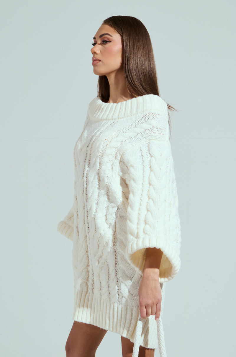A person with long hair is wearing the LALA LOVE YOU SWEATER DRESS, a chunky, white, off-the-shoulder cable-knit sweater with a large collar and wide sleeves. They stand against a plain background, looking to the side.