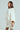 A person with long hair is wearing the LALA LOVE YOU SWEATER DRESS, a chunky, white, off-the-shoulder cable-knit sweater with a large collar and wide sleeves. They stand against a plain background, looking to the side.