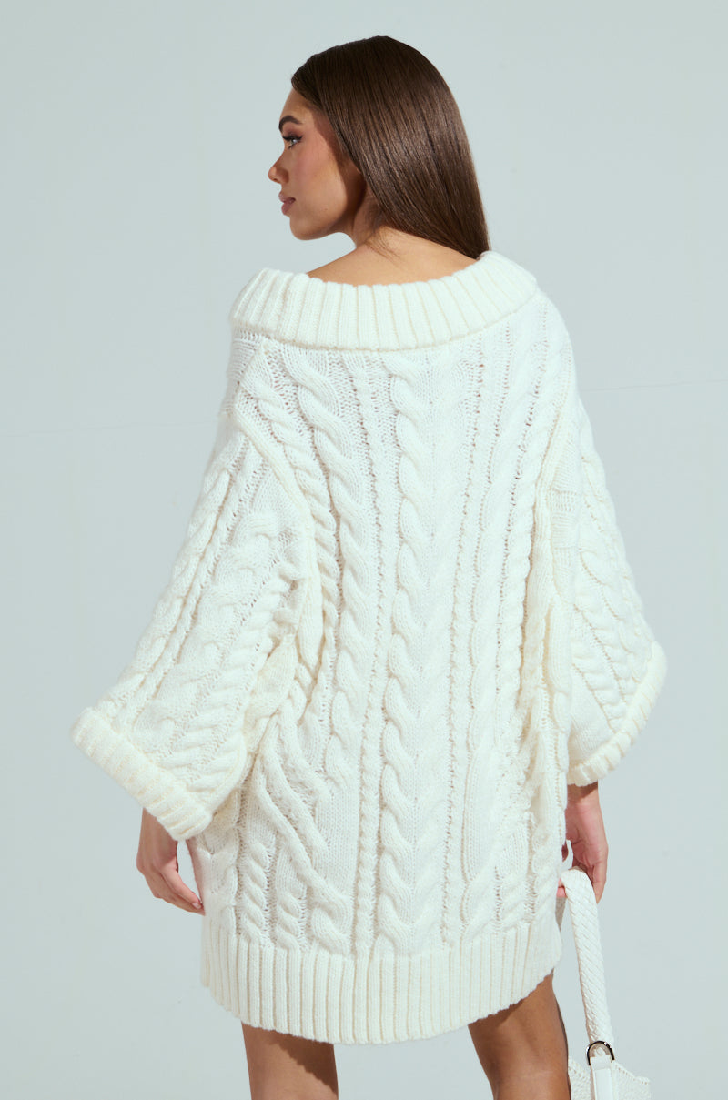 A woman in a LALA LOVE YOU oversized cable-knit sweater dress with an off-the-shoulder collar gazes sideways, carrying a white handbag. Her long, straight brown hair stands out against the pale blue background.