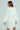 A woman in a LALA LOVE YOU oversized cable-knit sweater dress with an off-the-shoulder collar gazes sideways, carrying a white handbag. Her long, straight brown hair stands out against the pale blue background.