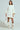 Against a plain backdrop, an individual models the off-the-shoulder "LALA LOVE YOU SWEATER DRESS," featuring chunky white cable-knit with three-quarter sleeves, paired with white slouchy knee-high boots and holding a large textured white bag.