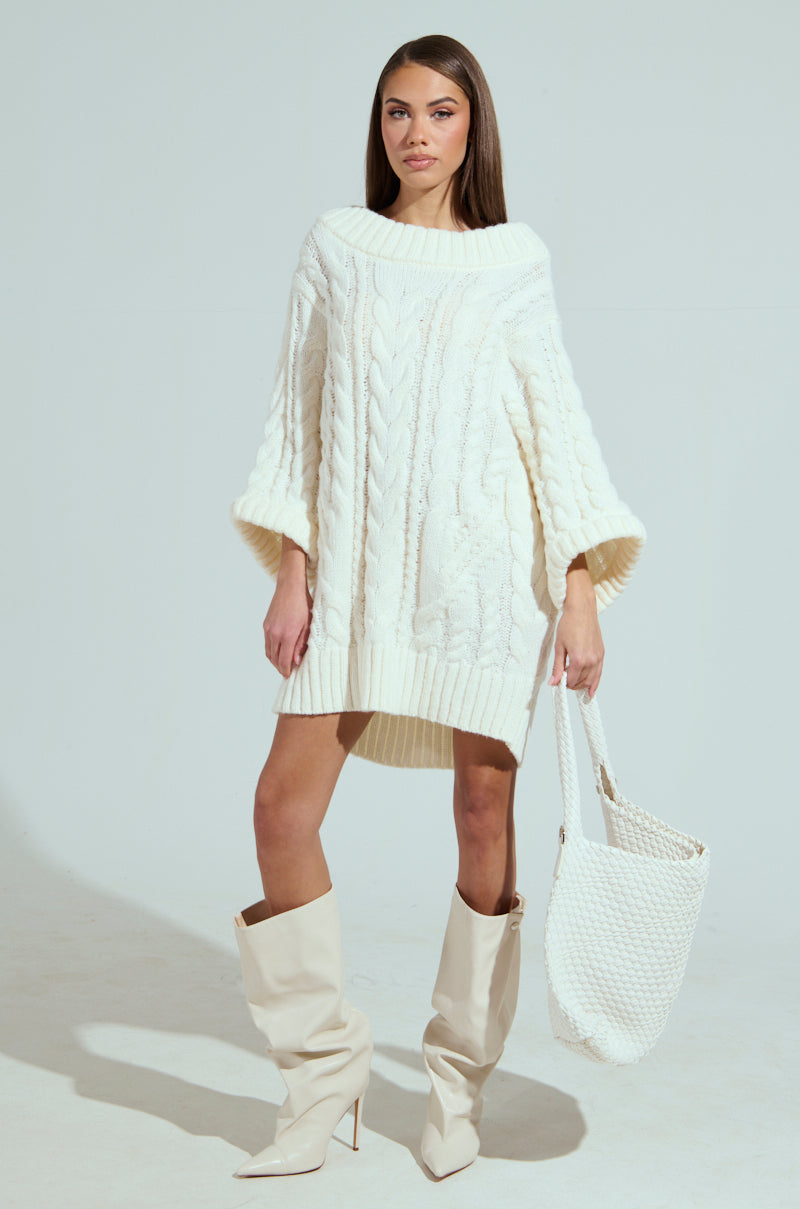A person confidently poses against a plain off-white background in an oversized off-the-shoulder cream LALA LOVE YOU SWEATER DRESS, complemented by matching knee-high cream boots and a white textured handbag.
