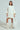A person confidently poses against a plain off-white background in an oversized off-the-shoulder cream LALA LOVE YOU SWEATER DRESS, complemented by matching knee-high cream boots and a white textured handbag.