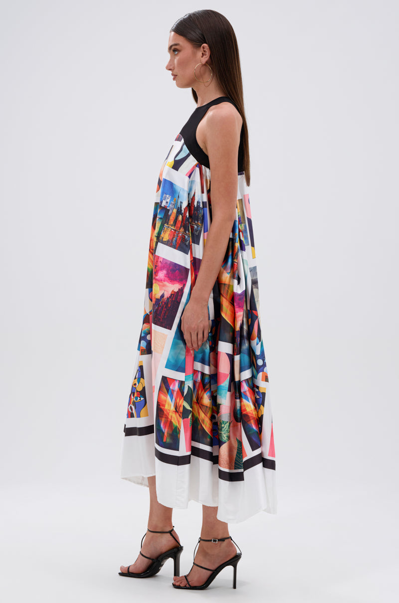 A woman stands sideways wearing the PUT ME IN A MUSEUM PRINTED MIDI DRESS, featuring a collage of colorful abstract prints and cityscapes. She has straight brown hair, black heeled sandals, and is set against a plain background.
