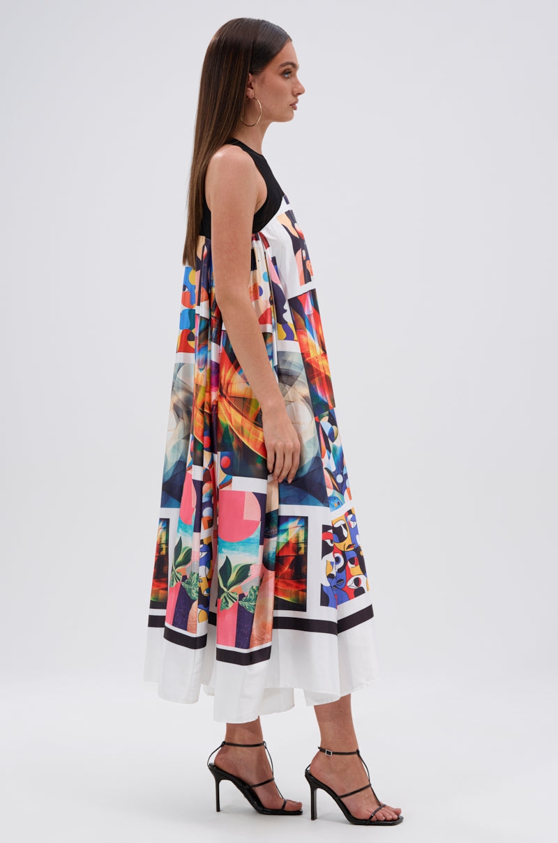 A woman wears the AKIRA Label PUT ME IN A MUSEUM PRINTED MIDI DRESS, a colorful geometric sleeveless dress with black straps, paired with black high-heeled sandals, standing sideways against a plain background.