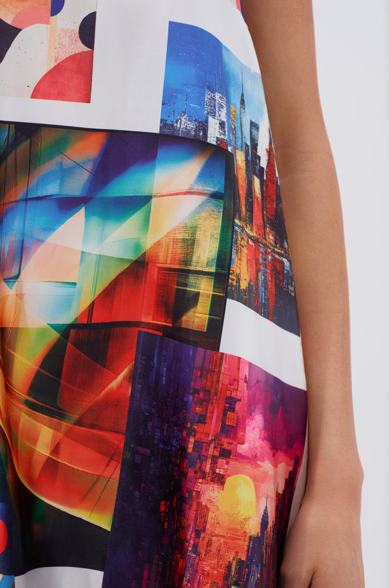 A close-up of the PUT ME IN A MUSEUM PRINTED MIDI DRESS, showcasing vibrant abstract cityscape prints in bold red, blue, orange, and yellow; a woman's arm and hand appear on the right.