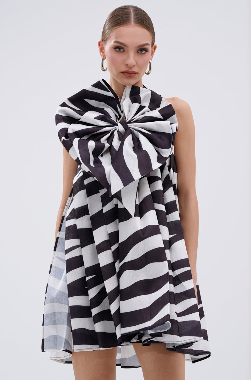 A woman wearing the WILD THING BOW FRONT MINI DRESS—a sleeveless mini with black-and-white zebra print and a large matching bow on one shoulder—stands against a plain background, confidently facing the camera.