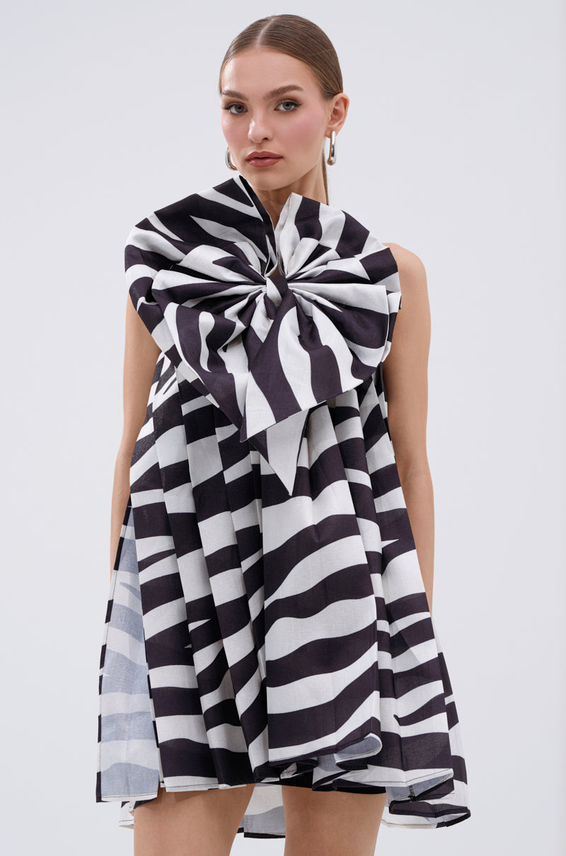 A woman with slicked-back hair and hoop earrings stands against a plain light background, wearing the WILD THING BOW FRONT MINI DRESS featuring a bold black-and-white zebra print and a large matching bow on the shoulder.
