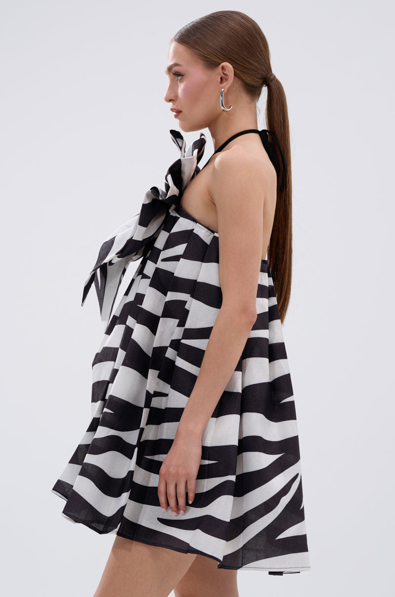 A woman in profile wears the WILD THING BOW FRONT MINI DRESS, showcasing a black and white zebra stripe pattern, oversized neckline bow, A-line silhouette, hoop earrings, and long straight hair tied back.