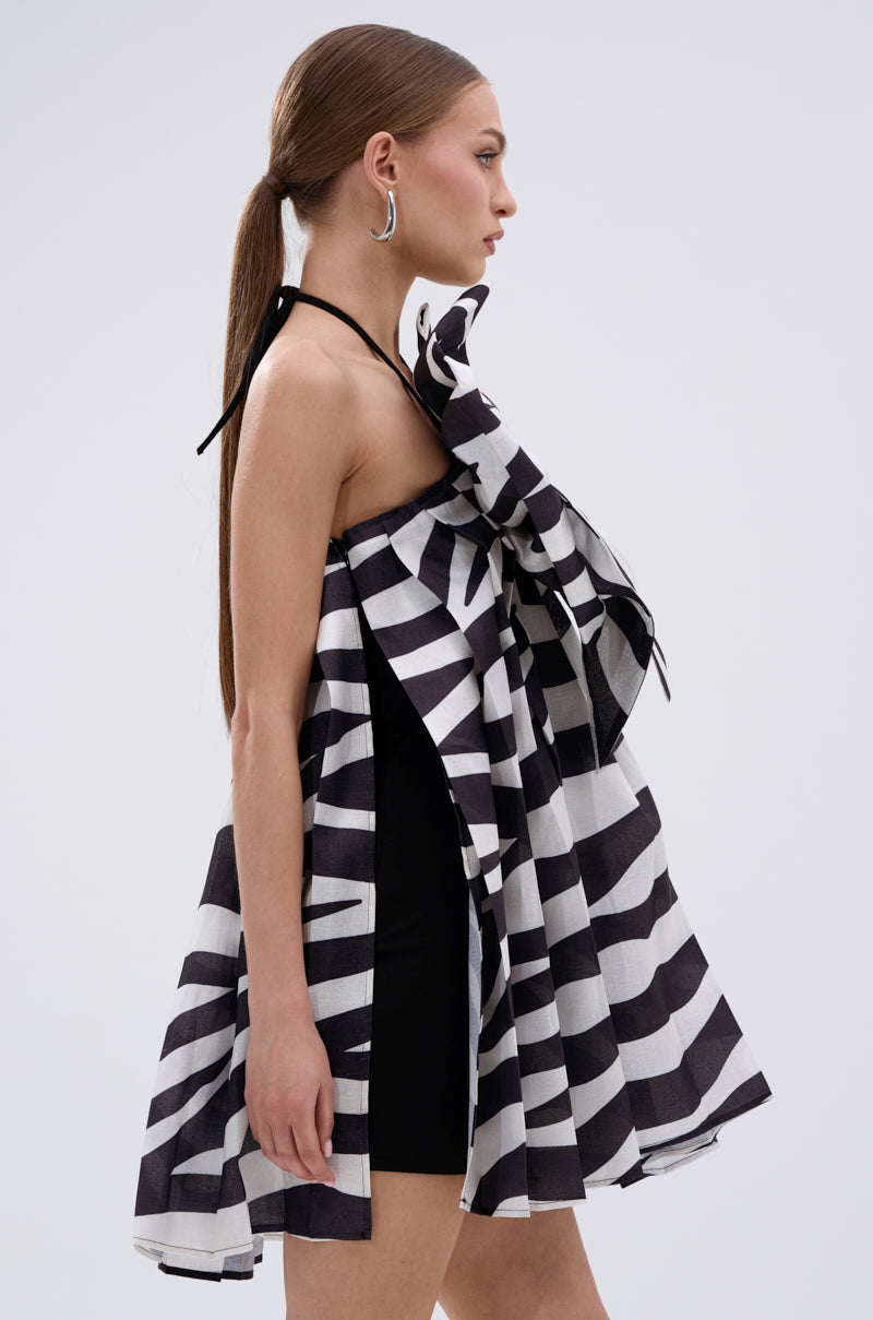 A woman in profile wears the WILD THING BOW FRONT MINI DRESS, a sleeveless black-and-white striped A-line mini with a large ruffled bow on the chest, black shorts underneath, silver hoop earrings, and her brown hair in a low ponytail.