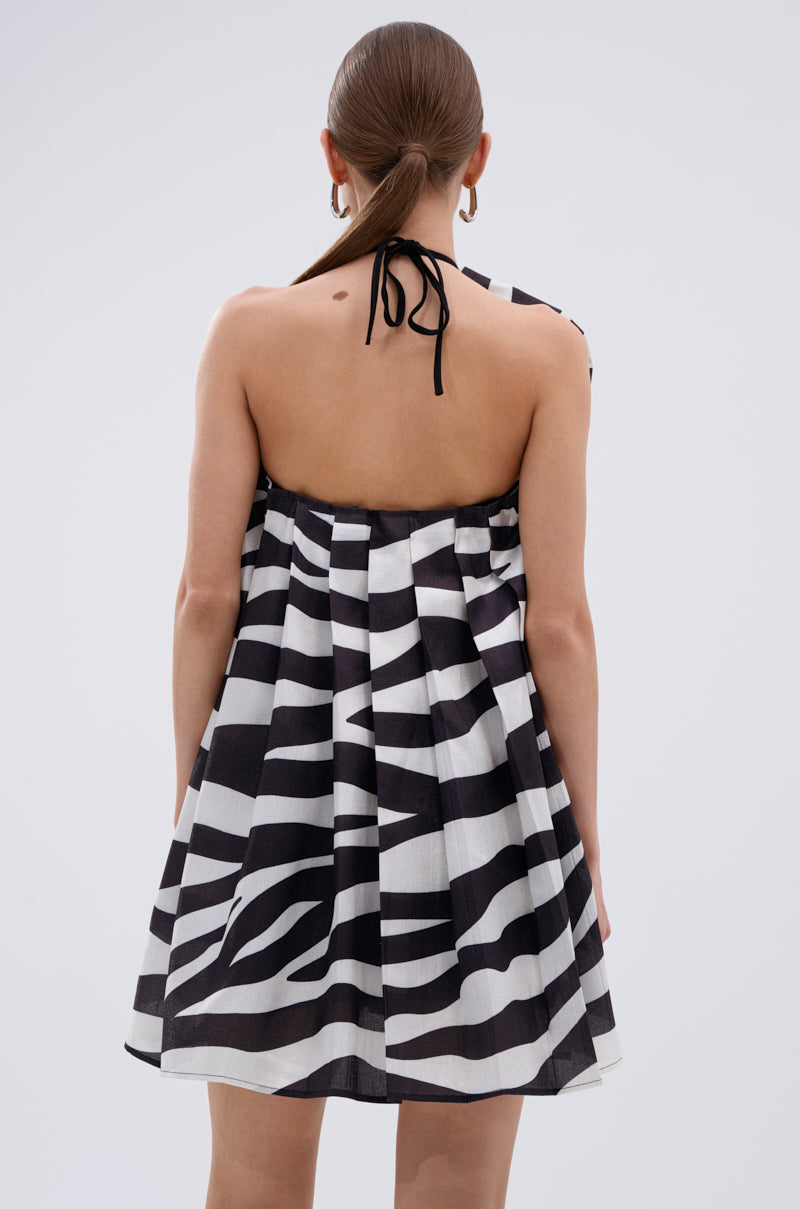 A person with straight brown hair tied back is wearing the WILD THING BOW FRONT MINI DRESS, showcasing a black and white abstract zebra print and halter tie-back. The photo is taken from behind against a plain light background.