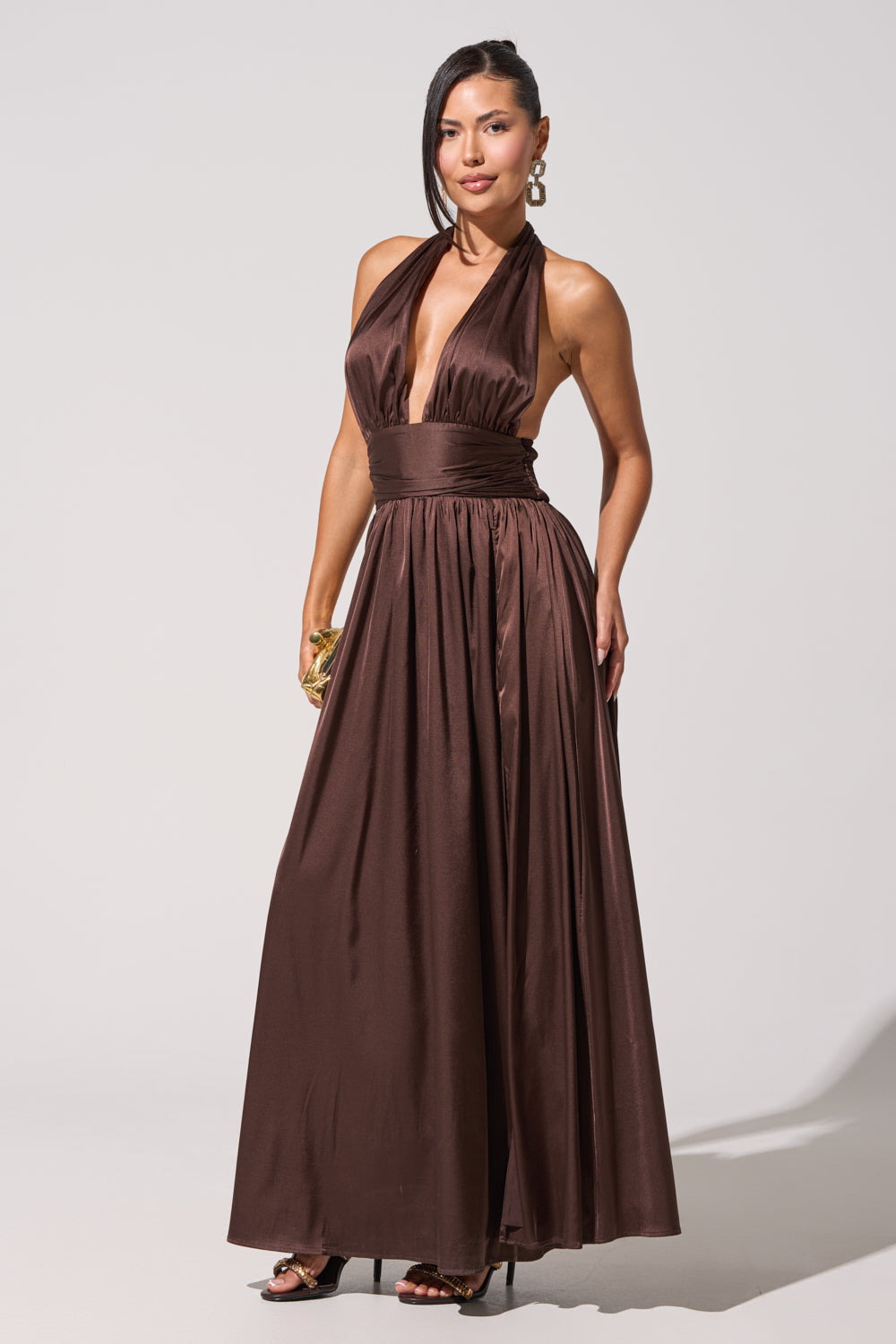 A woman poses confidently in the SET THE BAR MAXI DRESS, a brown satin halter maxi with a deep V-neck and wide waistband—styled with gold earrings, a bracelet, and high heels against a plain backdrop for an elegant wedding guest look. 3