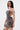 A woman with long straight hair stands sideways, looking at the camera, wearing the ISLAND GIRL PRINTED MINI DRESS—a fitted, sheer bodycon with a floral and abstract pattern. She holds a small chain bag and has a visible upper arm tattoo.