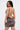 A woman with straight dark hair models the ISLAND GIRL PRINTED MINI DRESS, a backless, sleeveless mini with a floral print. Facing away and looking over her shoulder, she reveals tattoos on her arms against a plain light background.