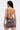 A person with long straight hair and hoop earrings stands facing away, showing off the backless, sleeveless ISLAND GIRL PRINTED MINI DRESS with a tropical floral pattern, visible arm tattoos, and holding a small chain-strap bag.