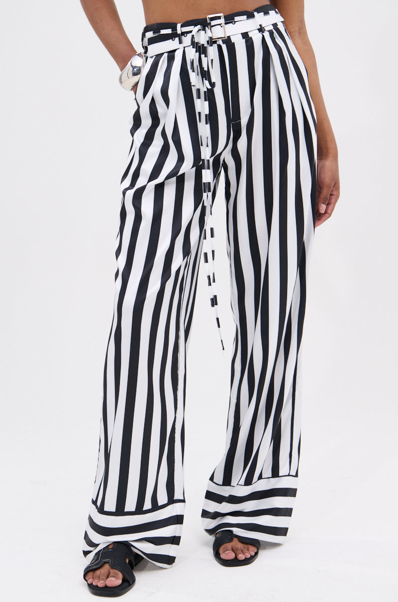 A person wearing BEETLE JUICE VIBES SATIN PANTS—a high-rise, wide-leg style with bold black and white stripes and a matching belt—paired with black sandals. The image is cropped from the torso to just above the sandals.