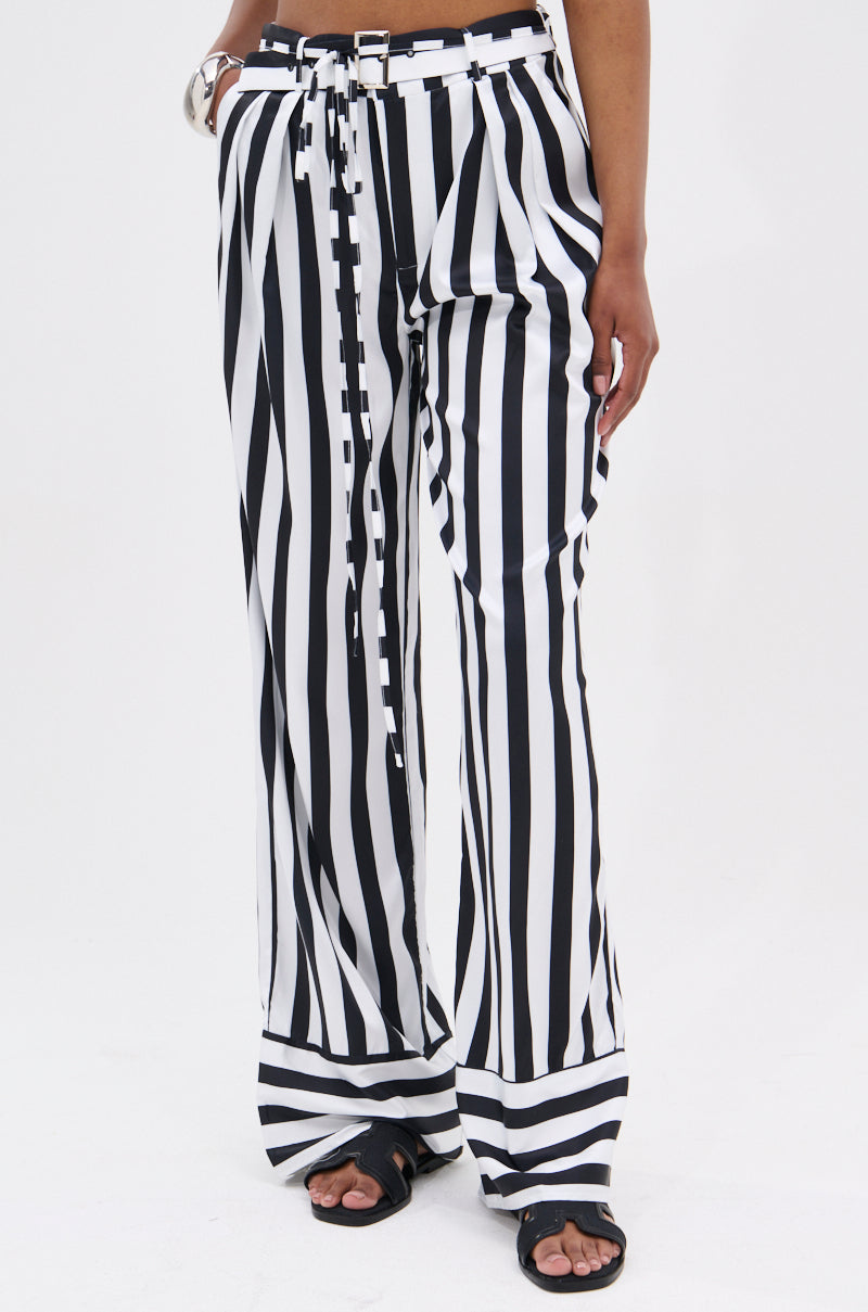 A person models the BEETLE JUICE VIBES SATIN PANTS—wide-leg, B&W striped satin with a matching belt and black sandals—standing against a white background. The cropped image highlights the pants' high-rise fit.