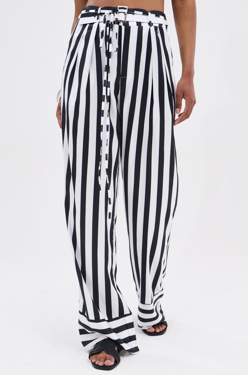 A person wearing BEETLE JUICE VIBES SATIN PANTS—white, high-rise with black stripes and a matching tie belt—paired with black sandals, stands against a plain white background.