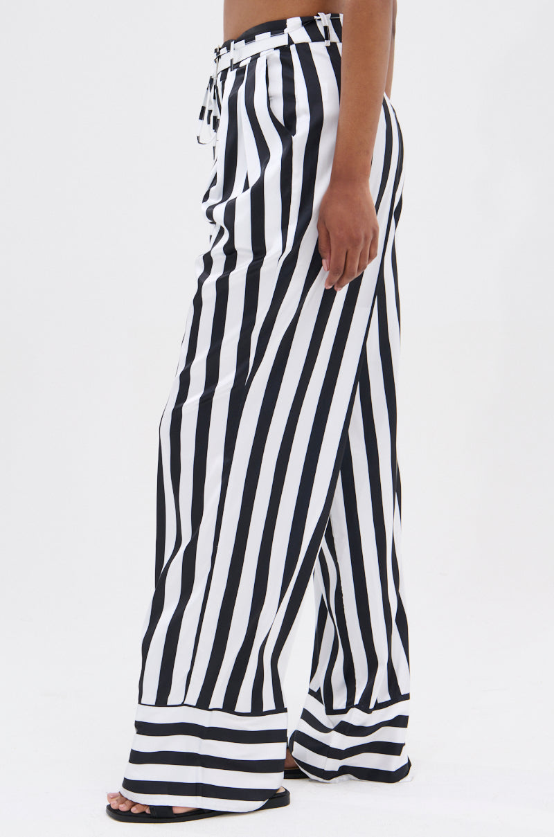 Someone in BEETLE JUICE VIBES SATIN PANTS—high-rise, wide-leg, black and white striped satin—pairs them with black sandals and stands in side profile against a plain white background.