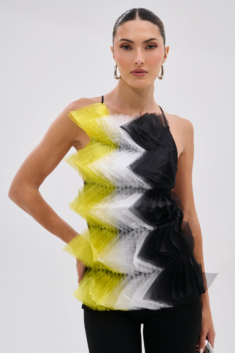 A woman wears the PIPPA MESH BLOUSE, an avant-garde top with zigzag layers of yellow, white, and black tulle ruffles, paired with black pants. She has slicked-back hair, hoop earrings, and poses against a plain light background.