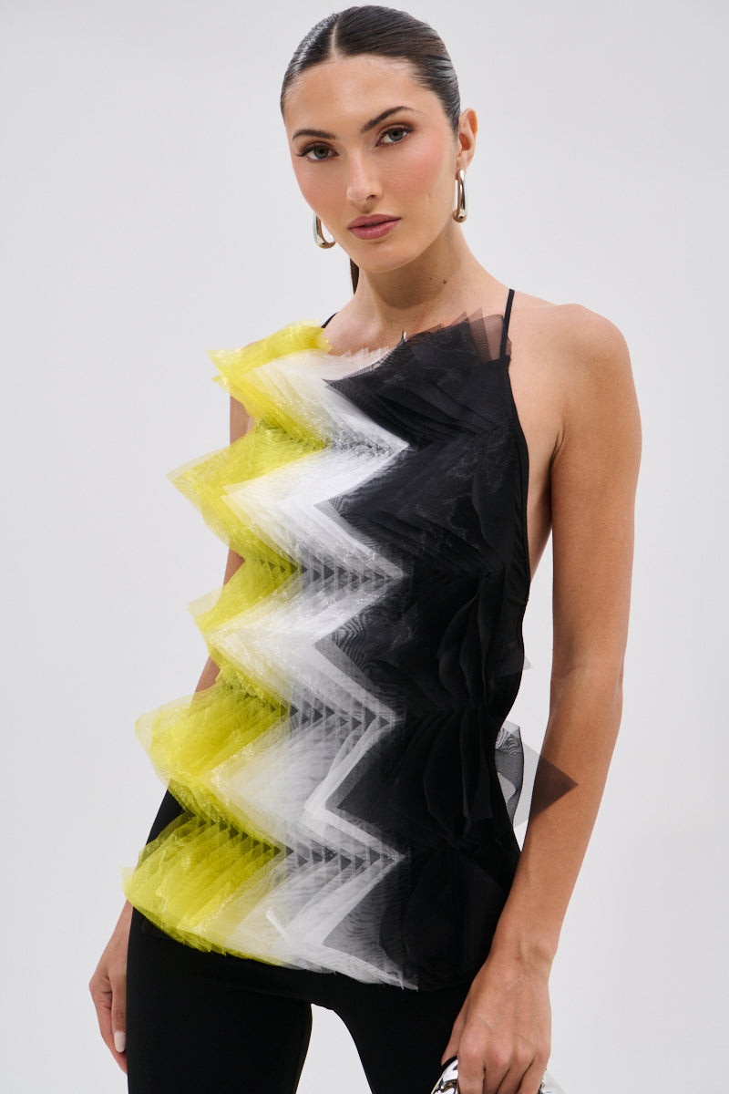A woman wearing the PIPPA MESH BLOUSE—an avant-garde sleeveless top with bold yellow, white, and black zigzag tulle layers—stands against a plain light backdrop. She has slicked-back hair, hoop earrings, and carries a small bag.